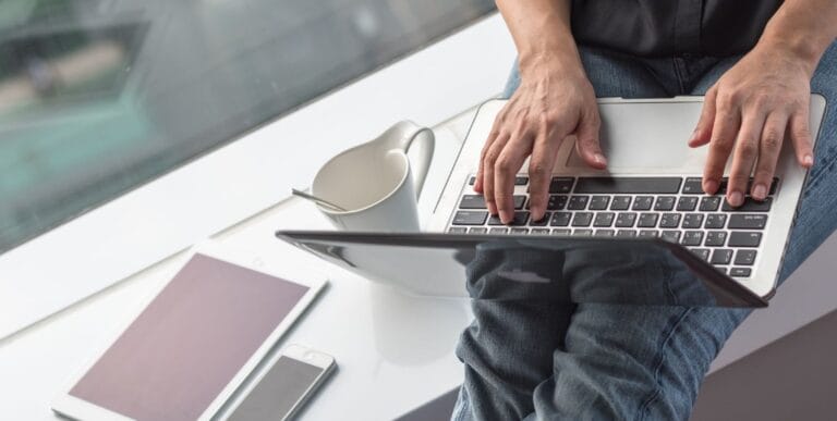 Mand sidder ved vindue med en kaffekop, mobiltelefon og tablet, mens han surfer på nettet med bærbar